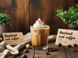 Hazelnut iced coffee topped with whipped cream and cinnamon, served in a glass with recipe cards, whole hazelnuts, coffee beans, and potted plants on rustic wooden table