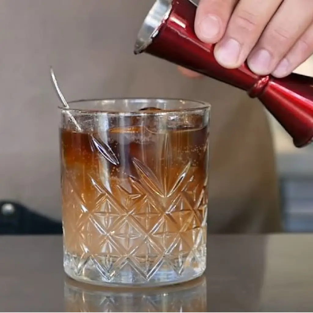 Cold Brew Tonic being prepared with hand pouring cold brew concentrate from red jigger into crystal glass with tonic water and ice