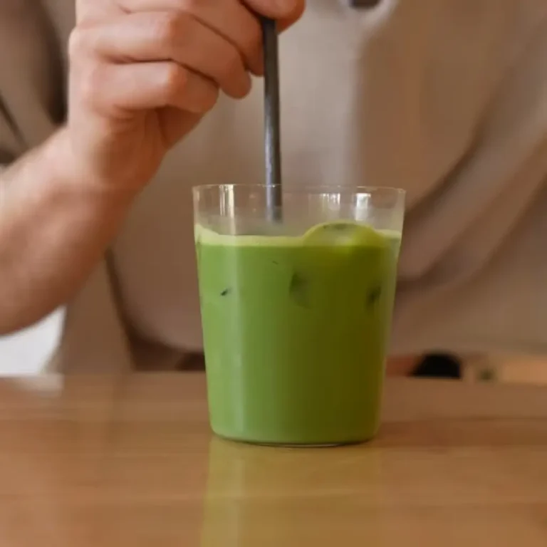 A hand stirring a refreshing homemade iced matcha latte in a clear glass with ice cubes on a wooden table, How to Make Iced Matcha Latte
