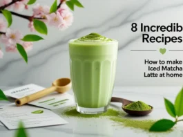 Creamy iced matcha latte in a tall glass with matcha powder, bamboo scoop, recipe cards, and cherry blossoms on marble surface☕ How to Make Iced Matcha Latte at Home💚☕