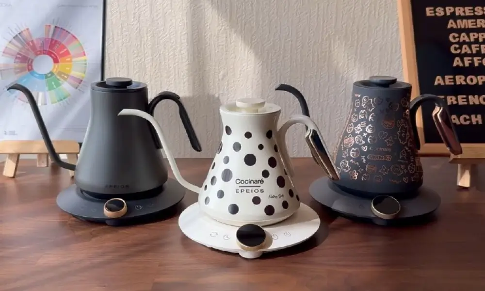 Three Cocinare Epeios gooseneck electric kettles displayed side by side on a walnut countertop β matte black, white polka dot, and dark illustrated editions β precision temperature control espresso accessories for the perfect manual coffee brewing ritual.