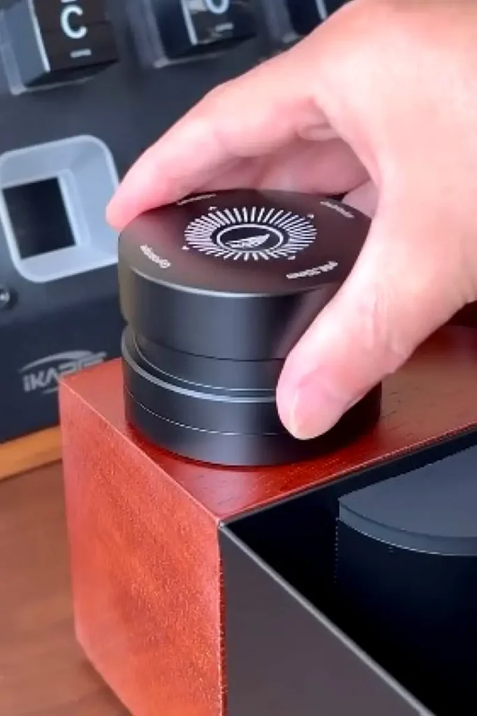 Hand holding the IKAPE Coffee Distributor 53.3mm in matte black with an engraved compass dial on top, resting on a walnut wood organizer station β a precision espresso distribution tool for leveling the coffee bed before tamping.