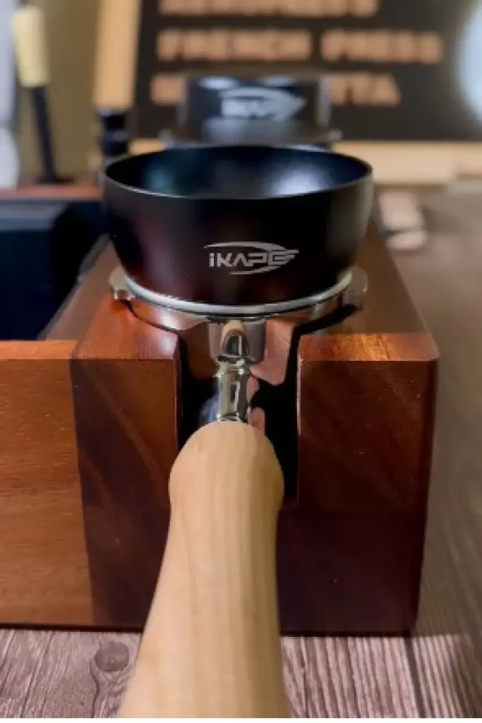 IKAPE Magnetic Dosing Funnel in matte black attached to a chrome portafilter with a wooden handle, resting in a walnut wood organizer station β an essential espresso distribution tool for clean, waste-free coffee dosing.