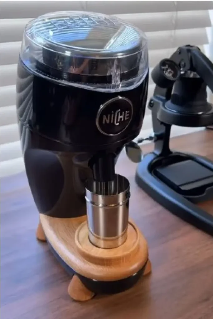 Niche Duo premium flat burr coffee grinder in matte black with a natural wood base and stainless steel dosing cup, positioned next to a manual espresso machine on a walnut countertop — a key espresso tool for precision home brewing.