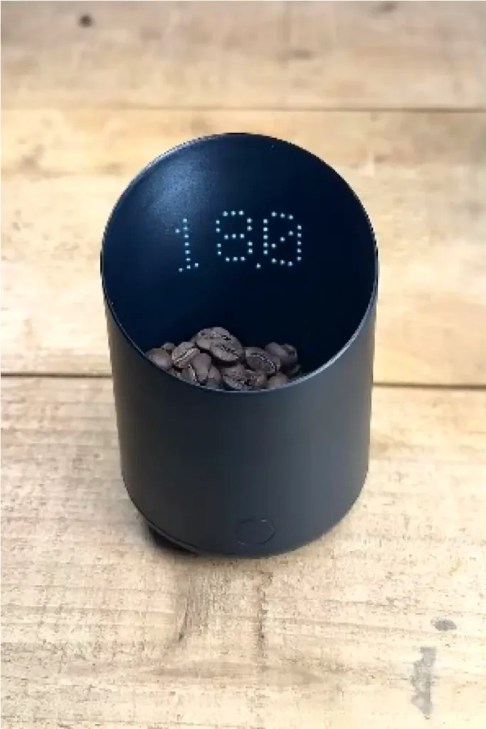 Subminimal Subscale digital dosing cup in matte black displaying an LED weight reading, filled with whole coffee beans on a rustic wooden surface β a precision espresso tool for accurate bean dosing.